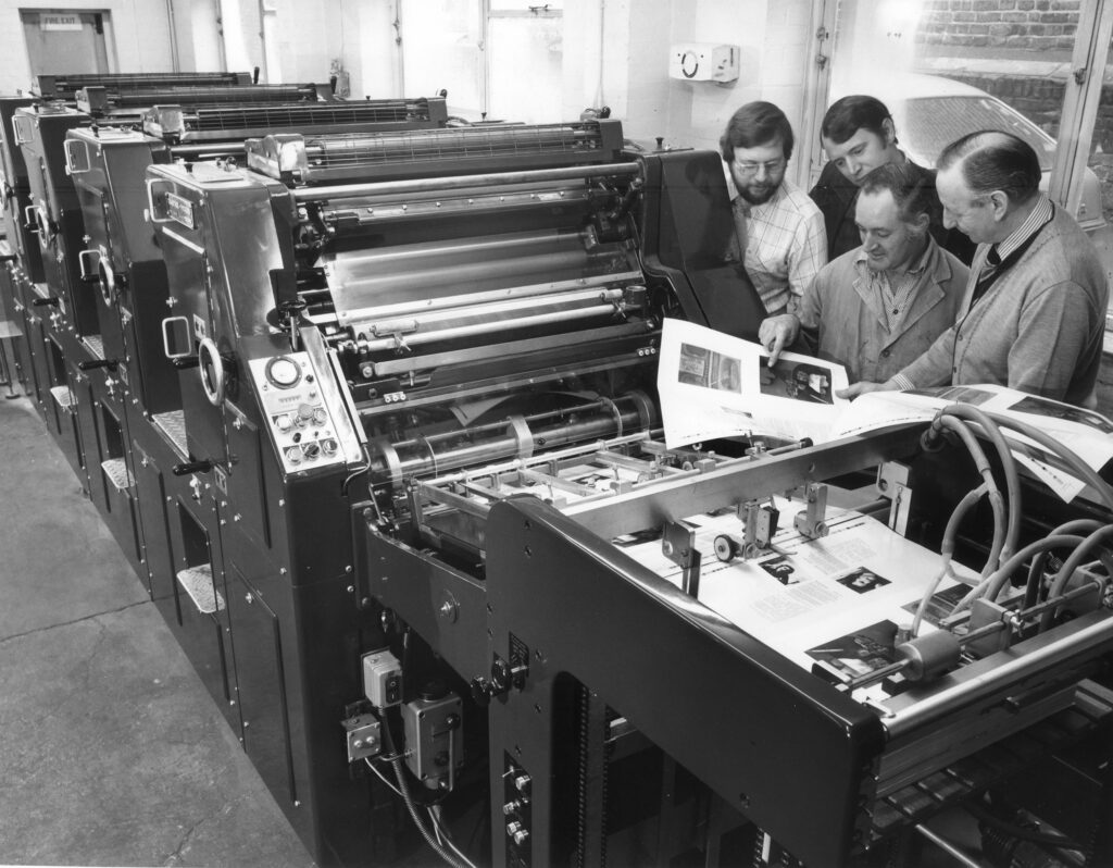 Four men looking at printed sheet by a printing press
